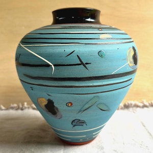 Könnte beinhalten: Eine bauchige Keramikvase mit matter türkisblauer Oberfläche, verziert mit horizontalen schwarzen und weißen Linien, verstreuten abstrakten Formen in Gelb, Schwarz und Weiß sowie einem glatten, dunkelbraunen kreisförmigen Rand an der Öffnung.