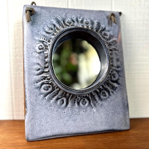 May include: A rectangular ceramic wall mirror with a light blue and grey speckled glaze. The frame features a circular center cutout surrounded by a textured, embossed pattern of small circles and radial lines. Two leather cords are attached at the top.