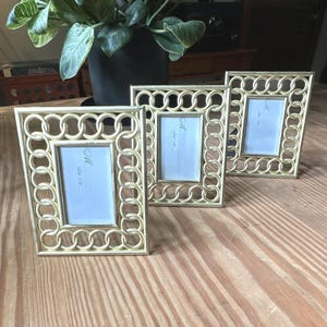 May include: Three rectangular picture frames with a metallic gold finish and a decorative border featuring a repeating interlocking circular chain link pattern. Each frame contains a white insert with the handwritten text M Table 04.
