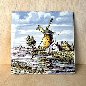 May include: A square ceramic tile featuring a hand-painted illustration of a Dutch landscape. The scene depicts a yellow windmill with rotating blades, a small white house with a dark roof, and a river flowing through grassy banks under a blue sky.