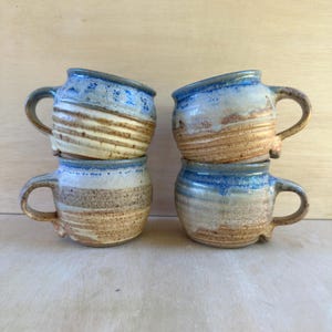 May include: Four ceramic mugs with rounded bodies and handles, featuring a two-tone glaze. The upper sections are a vibrant blue, while the lower portions display a textured, speckled tan and cream finish with visible horizontal ridges.