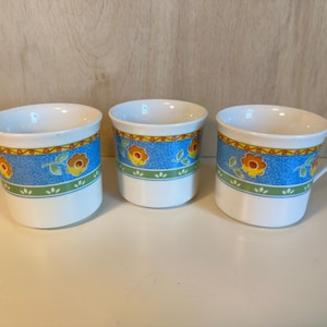May include: Three white ceramic mugs featuring a decorative band around the center. The band displays a blue textured background with orange and yellow floral patterns, green leaves, and a thin orange wavy line border on a white base.