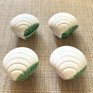 Könnte beinhalten: Vier weiße, muschelförmige Keramikschalen, die in einer quadratischen Formation angeordnet sind. Jede Schale hat eine glänzende Oberfläche mit konzentrischen Rillen und einen markanten grünen Akzent auf einer Seite, der das natürliche Aussehen einer Muschel nachahmt.
