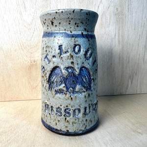 Op de afbeelding: Een cilindrische keramische steengoed mok met een gespikkelde lichtgrijze afwerking en blauwe accenten. De voorzijde toont een reliëf van een blauwe adelaar tussen de tekst ST. LOUIS en MISSOURI, met blauwe banden aan de boven- en onderrand.