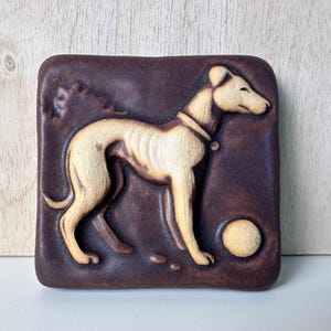 May include: A square ceramic tile featuring a raised relief of a cream-colored greyhound dog standing in profile. The dog wears a collar and faces a small circular ball. The background of the tile is a dark brown, matte-textured surface.
