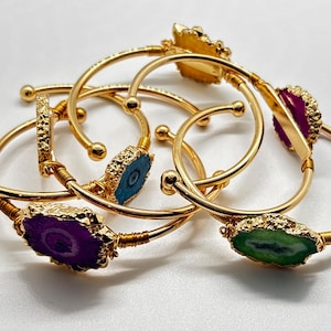 May include: A collection of gold-toned wire cuff bracelets, each featuring a single polished agate slice in vibrant colors including purple, green, blue, and magenta. The metal bands are thin and flexible, with small spheres at the open ends.