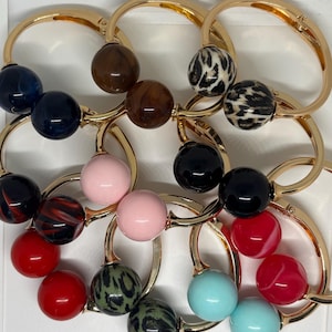 May include: A collection of circular gold-tone metal bangles, each featuring two spherical beads. The beads come in various finishes, including solid colours like red, pink, black, and light blue, as well as marbled and leopard print patterns.