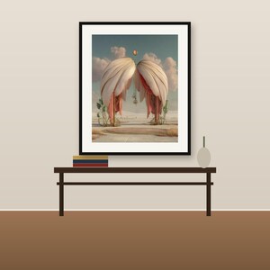 May include: A framed wall art print featuring a surreal landscape with two large, curved, cream-colored fabric structures resembling wings or drapes, set against a cloudy sky above a desert floor with a circular rug and sparse vegetation.