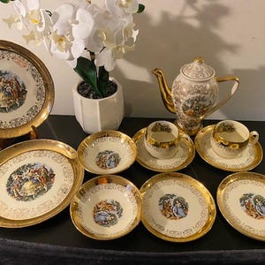 May include: A porcelain tea set comprising a teapot, two cups with saucers, two small bowls, and two plates. Each piece is cream-coloured with gold-toned rims and intricate filigree patterns, decorated with colourful illustrations of 18th-century figures.