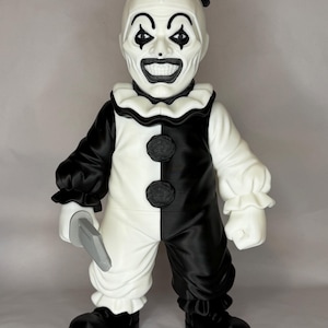 May include: A black and white clown figurine wearing a small top hat, a ruffled collar, and a two-toned jumpsuit with two circular buttons. The clown has a wide, toothy grin, dark eye makeup, and holds a grey toy knife in its right hand.