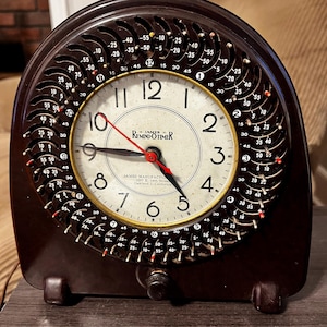May include: A dark brown James Remind-O-Timer clock with a cream-colored face, black numerals, and a red second hand. The outer ring features a circular dial with small metal pins and numerical markings for setting time-based reminders.