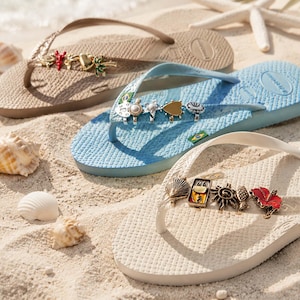 Op de afbeelding: Drie paar rubberen slippers in beige, lichtblauw en wit, elk met een getextureerd voetbed en een teenband versierd met diverse metalen bedels, waaronder hartjes, bloemen, schelpen en tropische motieven, geplaatst op een zanderige ondergrond.
