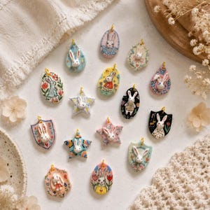 May include: A collection of fifteen small, decorative ceramic pendants featuring sculpted white rabbit faces. Each pendant has a unique shape, including stars, shields, and ovals, decorated with colourful hand-painted floral and patterned designs.