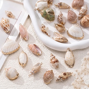 Può includere: Una collezione di conchiglie naturali dotate di tappi e anelli in metallo color oro per la creazione di gioielli. L'assortimento comprende varie conchiglie a spirale, coniche e a ventaglio in tonalità bianco, beige, marrone e verde.