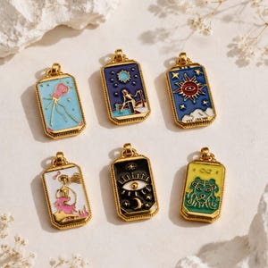 May include: Six rectangular gold-toned metal pendants featuring colourful enamel designs. The pendants display various illustrations including a pink figure, a seated person under stars, a sun, a person in a pink dress, an eye, and a green lion head.