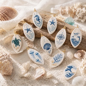 May include: A collection of ten oval-shaped white ceramic pendants featuring blue and gold nautical illustrations including a sea turtle, seahorse, starfish, compass, seashell, jellyfish, coral, dolphins, and a large wave, each with a gold jump ring.