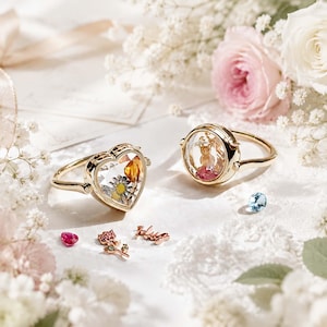 May include: Two gold-toned rings featuring clear glass locket centers containing small floral and decorative elements. One ring is heart-shaped, the other is circular. Nearby are loose charms including a pink gem, a rose, and the word "Hug".