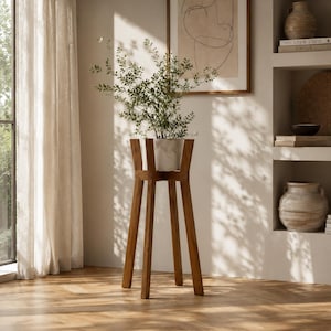 Op de afbeelding: Een hoge houten plantenstandaard met vier rechte poten en een vierkant bovenframe, met daarin een lichtgekleurde cilindrische pot met een groene bladplant met dunne takken. De standaard is gemaakt van hout in een middelbruine tint.