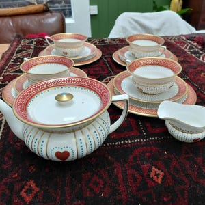 Peut inclure: Un service à thé comprenant une théière, un pot à lait et quatre tasses avec soucoupes assorties. Les pièces en porcelaine blanche sont ornées de motifs de cœurs rouges, de lignes verticales pointillées bleues et de bordures dorées.