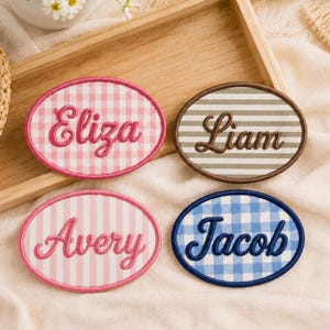 May include: Four oval fabric patches with embroidered names. One pink gingham patch reads Eliza, one brown striped patch reads Liam, one pink striped patch reads Avery, and one blue gingham patch reads Jacob. Each patch has a stitched border.