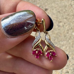 May include: A pair of drop earrings featuring a gold-tone metal frame with a twisted design. Each earring is accented with a small row of clear stones and three marquise-cut, deep pink-red stones arranged in a cluster at the bottom of the drop.