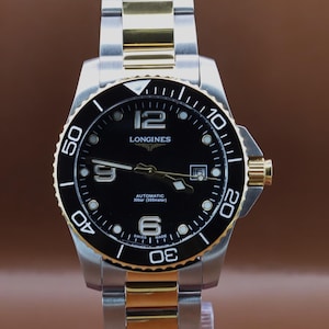 May include: A two-tone stainless steel and gold-tone Longines automatic watch with a black dial, featuring a rotating bezel with white numerals, a date window at the three o'clock position, and a metal link bracelet with a folding clasp mechanism.
