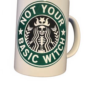 May include: A white ceramic mug featuring a circular green and black graphic on the front. The design depicts a skeleton figure with long hair and a crown, surrounded by the text NOT YOUR BASIC WITCH in a bold, circular green border.