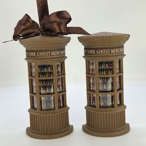 Op de afbeelding: Twee bruine cilindervormige ornamenten met een miniatuurwinkeldisplay met planken vol kleine kleurrijke figuren. Elk stuk is voorzien van de tekst THE YORK GHOST MERCHANT op de bovenrand. Eén ornament heeft een bruin satijnen lint.