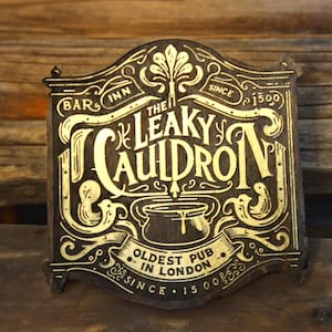 May include: A dark brown wooden sign featuring gold-colored engraved text and illustrations. The sign reads THE LEAKY CAULDRON, BAR INN SINCE 1500, and OLDEST PUB IN LONDON SINCE 1500. A central graphic depicts a cauldron with liquid dripping from the rim.