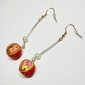 May include: A pair of gold-tone drop earrings featuring long, thin metal wires, each accented with a single white faux pearl and a spherical fabric charm. The fabric spheres display intricate floral patterns in shades of red, orange, and yellow.