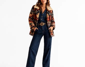 Op de afbeelding: Een donkerblauwe spijkerjumpsuit met een knoopsluiting aan de voorzijde en een bruine leren riem met een grote gesp. De jumpsuit wordt gedragen onder een jas met lange mouwen met een geometrisch patroon in rood, zwart en beige, afgewerkt met een crèmekleurige kraag.