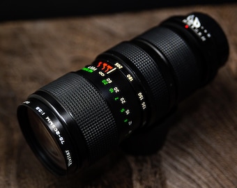May include: Black Vivitar 75-205mm f/3.8 camera lens. The lens has a textured grip and markings in green, white, and red. The lens has measurements in feet and millimeters. The lens is on a wooden surface.