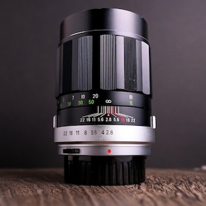 Puede incluir: Un objetivo de cámara negro y plateado con un diseño de barril acanalado. El objetivo tiene varias marcas, incluyendo números y escalas para el enfoque y la apertura. El objetivo está sobre un fondo oscuro.