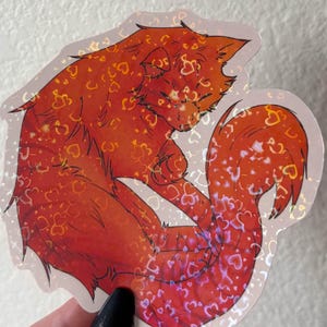 Puede incluir: Una pegatina troquelada que muestra un gato naranja enroscado con un acabado hologr&aacute;fico. El pelaje del gato est&aacute; cubierto por un patr&oacute;n repetitivo de peque&ntilde;os corazones brillantes que captan la luz, creando un efecto reflectante.