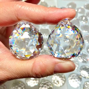 May include: Two clear, faceted crystal spheres with a teardrop shape. Each crystal features a geometric pattern of triangular cuts that refract light, creating a spectrum of rainbow colors across the surfaces of the transparent glass objects.