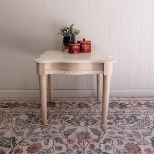 May include: A light-colored wooden side table with four tapered legs and a scalloped edge tabletop. The table features a carved decorative trim along the apron. On top sits a dark blue planter with a green plant and two red ribbed canisters.