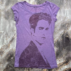May include: A light purple short-sleeved t-shirt featuring a dark purple distressed graphic print of a person's face and upper torso. The shirt has a round neckline and a fitted silhouette, with the graphic occupying the right side of the front.
