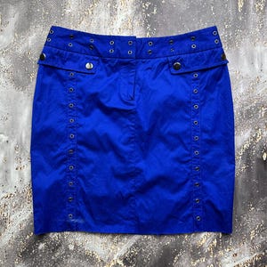 May include: A vibrant royal blue skirt featuring a structured waistband with multiple silver-toned metal eyelets. The front includes two decorative flap pockets with silver buttons and two vertical rows of eyelets running down the front panels.