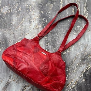 Puede incluir: Un bolso de hombro de color rojo brillante construido con varios parches de cuero de diferentes texturas, incluyendo patrones lisos y grabados. El bolso cuenta con dos correas largas para el hombro y una pequeña etiqueta de marca rectangular plateada con el nombre SILVANO.