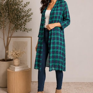 Pode incluir: Um cardigã longo e aberto com padrão xadrez verde e azul-marinho, mangas três quartos e tecido leve semitransparente. A peça é usada sobre uma regata branca simples, calças jeans azul-escuras e botas de cano curto na cor bege.