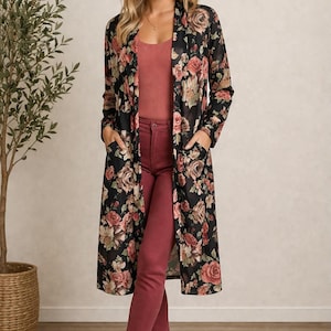 May include: A long-sleeved, open-front duster cardigan in black featuring a floral print with shades of pink, red, and green. The garment has two front patch pockets and falls to the knee, worn over a solid mauve tank top and matching pants.
