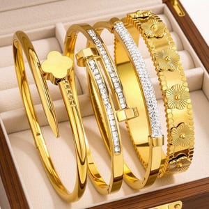 May include: A set of four gold-toned bracelets. One features a clover-shaped end, another is a nail-inspired design, the third is a band encrusted with clear stones, and the fourth is a wide band decorated with a series of raised floral motifs.