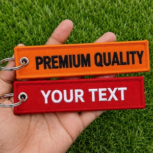Puede incluir: Dos llaveros rectangulares bordados con anillas metálicas. El llavero superior es naranja con el texto bordado en negro PREMIUM QUALITY. El llavero inferior es rojo con el texto bordado en blanco YOUR TEXT.