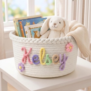 May include: A white woven rope storage basket featuring a braided top rim and the name Kelsey spelled out in colorful yarn letters. The basket is decorated with two small pink and purple yarn flowers and contains several books and a plush rabbit toy.