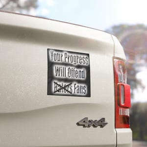 Op de afbeelding: Een vierkante bumpersticker met een zwart-witte gradiëntachtergrond en de tekst Your Progress Will Offend Haters Fans, waarbij het woord Haters is doorgehaald met een zwarte X, aangebracht op de achterkant van een wit voertuig met 4x4-embleem.