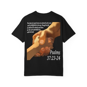 May include: A black short-sleeve t-shirt featuring a graphic of two hands holding each other. The text reads: The steps of a good man are ordered by the Lord: and he delighteth in his way. Though he fall, he shall not be utterly cast down: for the Lord upholdeth him with his hand. Psalms 37:23-24.