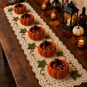 May include: A long, cream-colored crocheted table runner featuring an intricate lace border and decorative green crocheted leaves and vines. Five orange crocheted pumpkin-shaped bowls are placed along the runner, each filled with small candy pieces.
