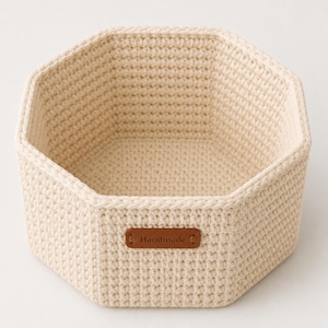 May include: An off-white, octagonal crochet basket made from thick cord. The basket features a textured, woven stitch pattern throughout its structure and includes a small, rectangular brown leather-like tag attached to the front with the word Handmade.