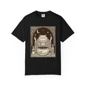 Puede incluir: Una camiseta negra de manga corta con un estampado gráfico de una rana con túnica, de pie detrás de un caldero humeante con dos botellas de vidrio a los lados, sobre un fondo oscuro con una luna creciente y estrellas.