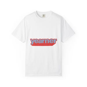 Può includere: Una t-shirt bianca a maniche corte con la parola "yearner" stampata in lettere minuscole azzurre al centro del petto. Il testo è posizionato su uno sfondo rettangolare rosso a tinta unita con angoli arrotondati, creando un design grafico audace.
